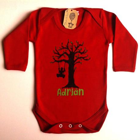 Body rojo para bebé con un árbol que tiene un columpio y un niño columpiándose. Todo en color negro excepto unas pequeñas hojas que nacen de las ramas y el nombre del niño que van en verdes