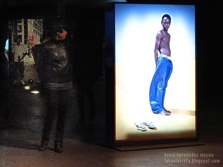 Una mujer mira en la oscuridad una fotografía de la exposición de Isabel Muñoz en la exposición de Caixa Forum, mientras el adolescente de la fotografía parece mirarla a ella en un intercambio de miradas