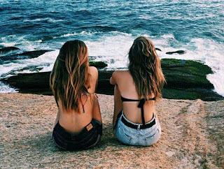 Dos amigas están sentadas juntas en una roca delante del mar, dando la espalda al fotógrafo. Ambas sin camiseta, con vaqueros cortos, uno azul y otro negro.