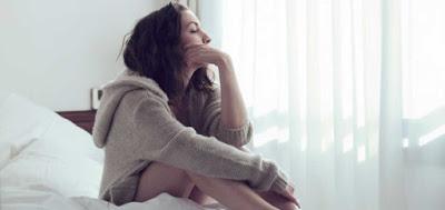 Mujer joven sentada en la cama, pensando, con una sudadera beige con gorro.