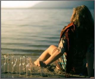 Chica joven sentada en la playa, mirando al mar, mientras que agarra una botella de cristal vacía, junto a muchas más.