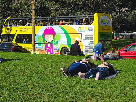 Sobre el césped gente recostada al sol y al fondo turista en bus esperando.