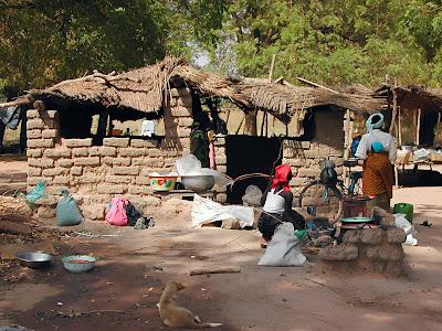 Fotografía real de la vida cotidiana en un mercado local de Burkina Faso, que muestra estructuras rústicas de adobe con techos de paja, personas interactuando en sus puestos y un perro descansando en primer plano sobre el suelo de tierra.