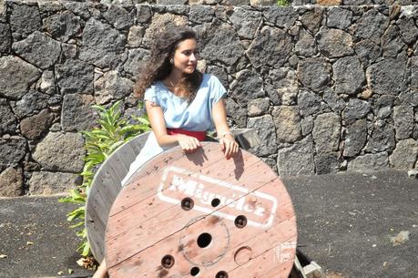 las fotos están hechas en lanzarote en el pueblo de masdache, podemos ver como silvia está vestida con un vestido vaquero con un cinturón fucsia que le bordea la cintura y unas sandalias planas de color camuflaje con plumas. las fotos están hechas sobre un barril de madera y árboles frutales a su alrededor
