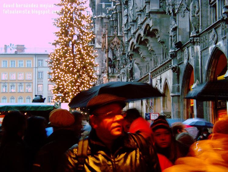 Escena de gente caminando en la plaza central de Municht, Alemania, mientras está lloviendo, con un mercadillo navideño y un árbol decorado con luces