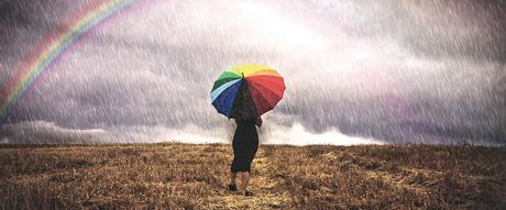 Una mujer con un paraguas camina bajo la lluvia en un campo, al fondo se ve un arco iris. Foto OMM