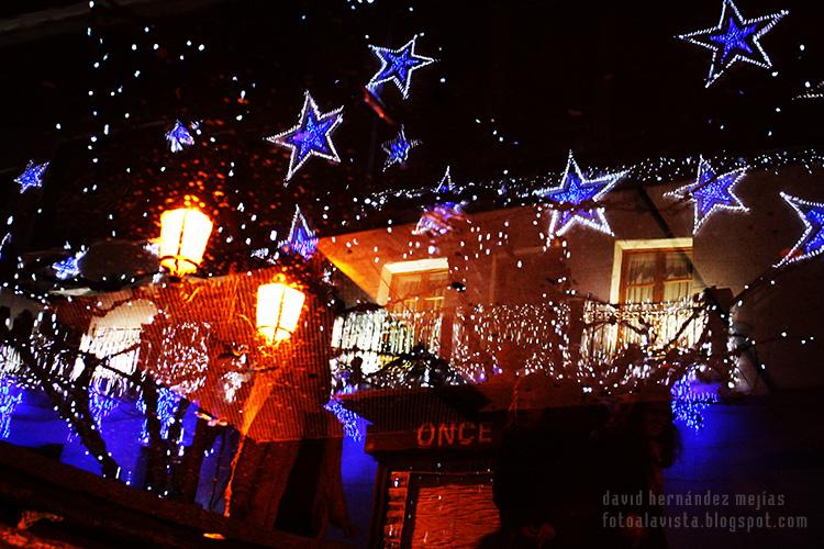 Estrellas luminosas y demás luces reflejadas junto a tres figuras en el agua nocturna de una pileta en la Plaza Mayor de Torrejón de Ardoz, Madrid (España). Fotografía inversa