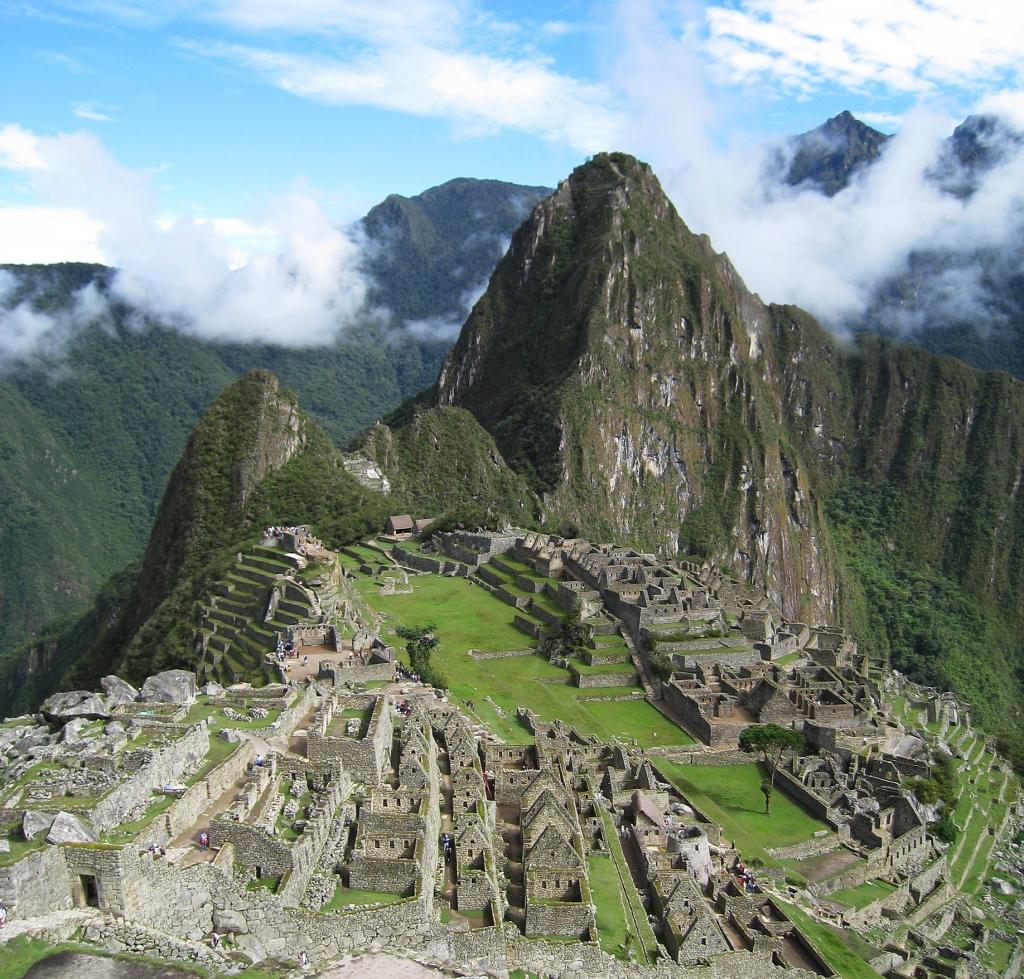 Vista de sur a norte. A la izquierda se ve el sector Hanan de la ciudad (con la estructura piramidal de la colina del Intihuatana) y a la derecha el sector Oriente, separadas por la plaza principal. Al fondo el Cerro Huayna Picchu. La imagen está tomada desde lo alto del sector agrícola, al sur del complejo. Wikipedia.