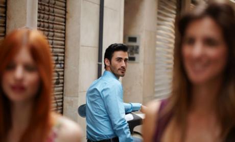 Chico joven con camisa azul se queda mirando a dos mujeres jóvenes, una castaña y otra pelirroja, mientras caminan.
