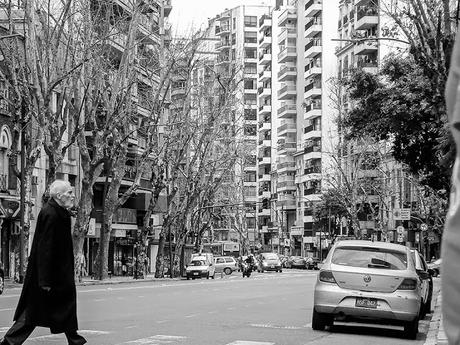 Hombre con sobretodo negro cruzando la avenida ,árboles y condominios al fondo.