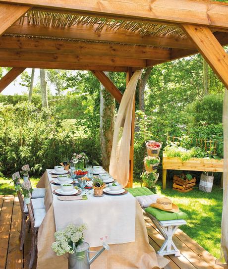 Comedor bajo pérgola de madera y cañas, con cortinas y manteles en tostados, blanco y beige, junto a jardín con huerto urbano 345652