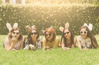 Cinco mujeres jóvenes y amigas tumbadas en el césped en un día soleado, todas mirando a la cámara con gafas de sol.