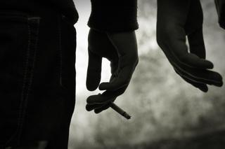 Manos de dos personas diferentes, la mano de la izquierda sostiene un cigarro mientras que la mano de la derecha, nada. Fondo gris e imagen oscura.