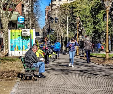 Señor leyendo en banco de plaza y joven caminando con celu en manos.