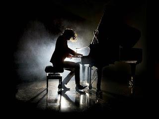 En la oscuridad, un hombre con el pelo largo toca el piano. Un foco al fondo ilumina desde atrás, por lo que solo se ve la silueta del piano y del pianista.