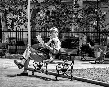 Un hombre sentado en banco de plaza al sol leyendo el diario.