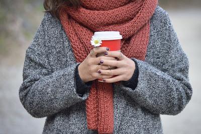 Mujer con jersey de lana, abrigo y bufanda gruesa, sosteniendo una taza de café y una margarita