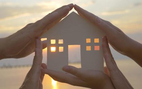 Group of human palms on all sides of a cut out house diagram with setting sun coming through window over water