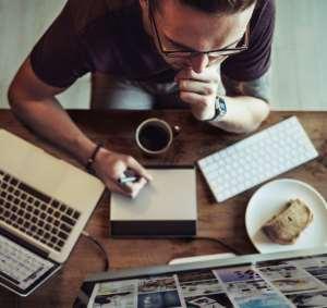 Un hombre en actitud pensativa sentado frente a un ordenador. A un lado tiene un cuaderno y un lápiz, una taza de café y un pastel de manzana.