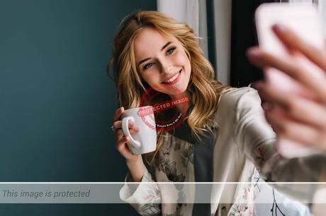 pretty-young-woman-with-wavy-blonde-hair-taking-selfie-sitting-window-with-cup-morning-coffee-tea-she-wearing-silk-pajama-turquoise-wall-min