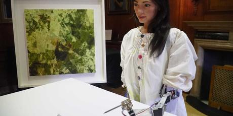 Foto de Ai-Da, el robot pintor, con una pintura en el fondo