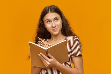 Creative teenage girl dressed in striped top holding diary and looking up