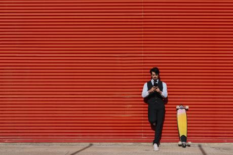 Male entrepreneur using mobile phone by longboard against wall