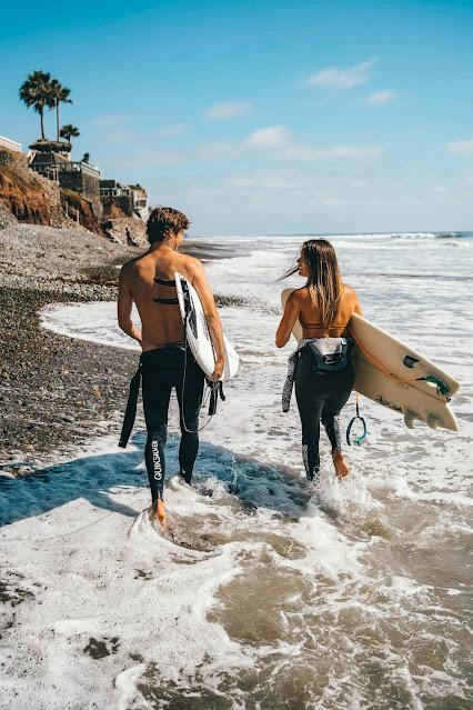 Pareja paseando por la orilla de la playa llevando tablas de surf